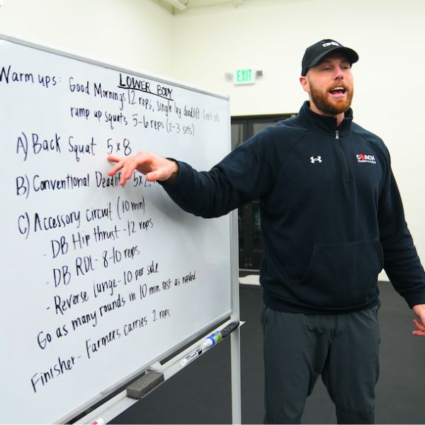 Man in athletic wear pointing at a whiteboard with workout instructions outdoors.